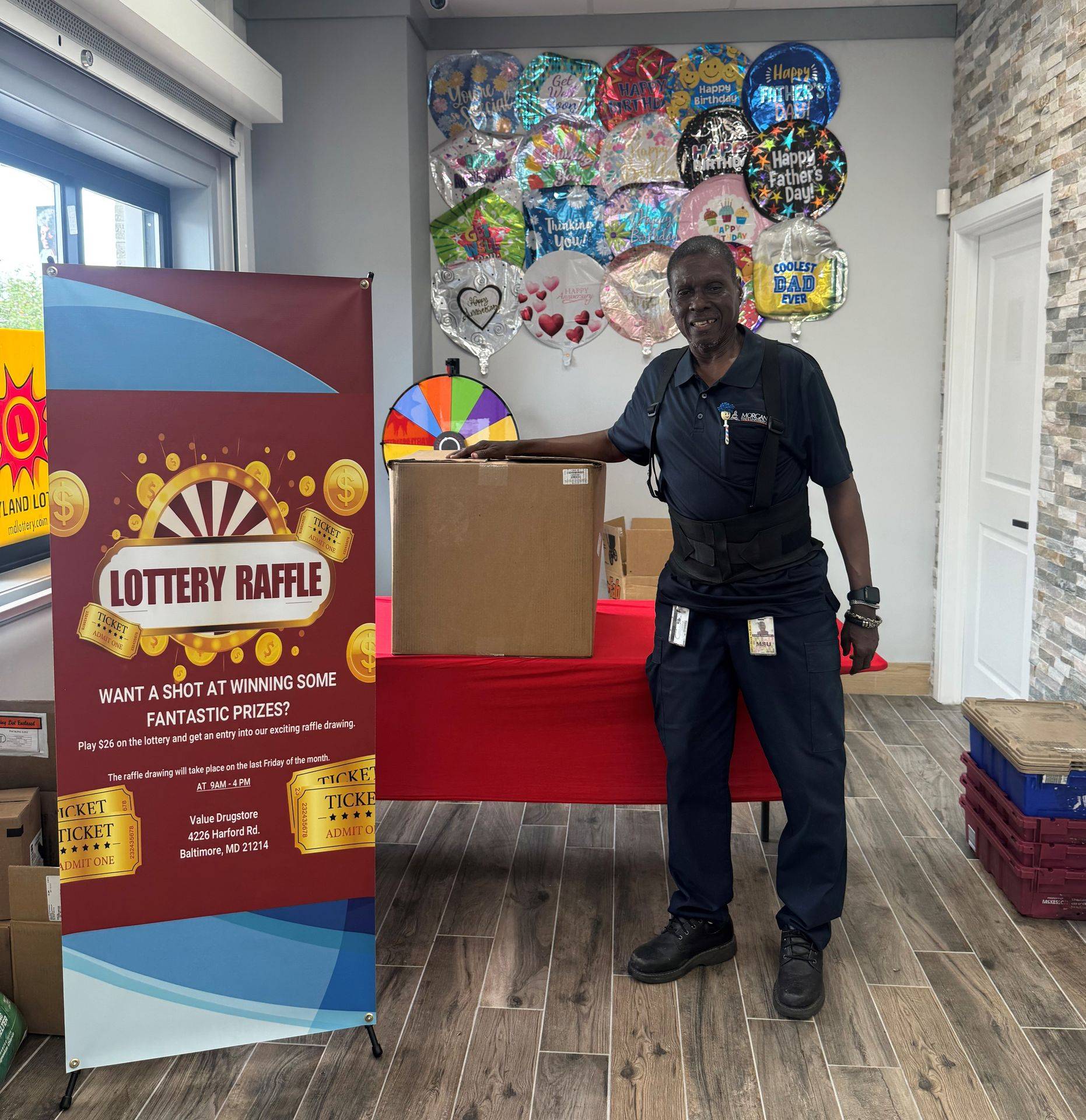 Lottery raffle banner with text promoting a chance to win prizes, a man in uniform standing next to a box, colorful balloons in the background, and a red table.