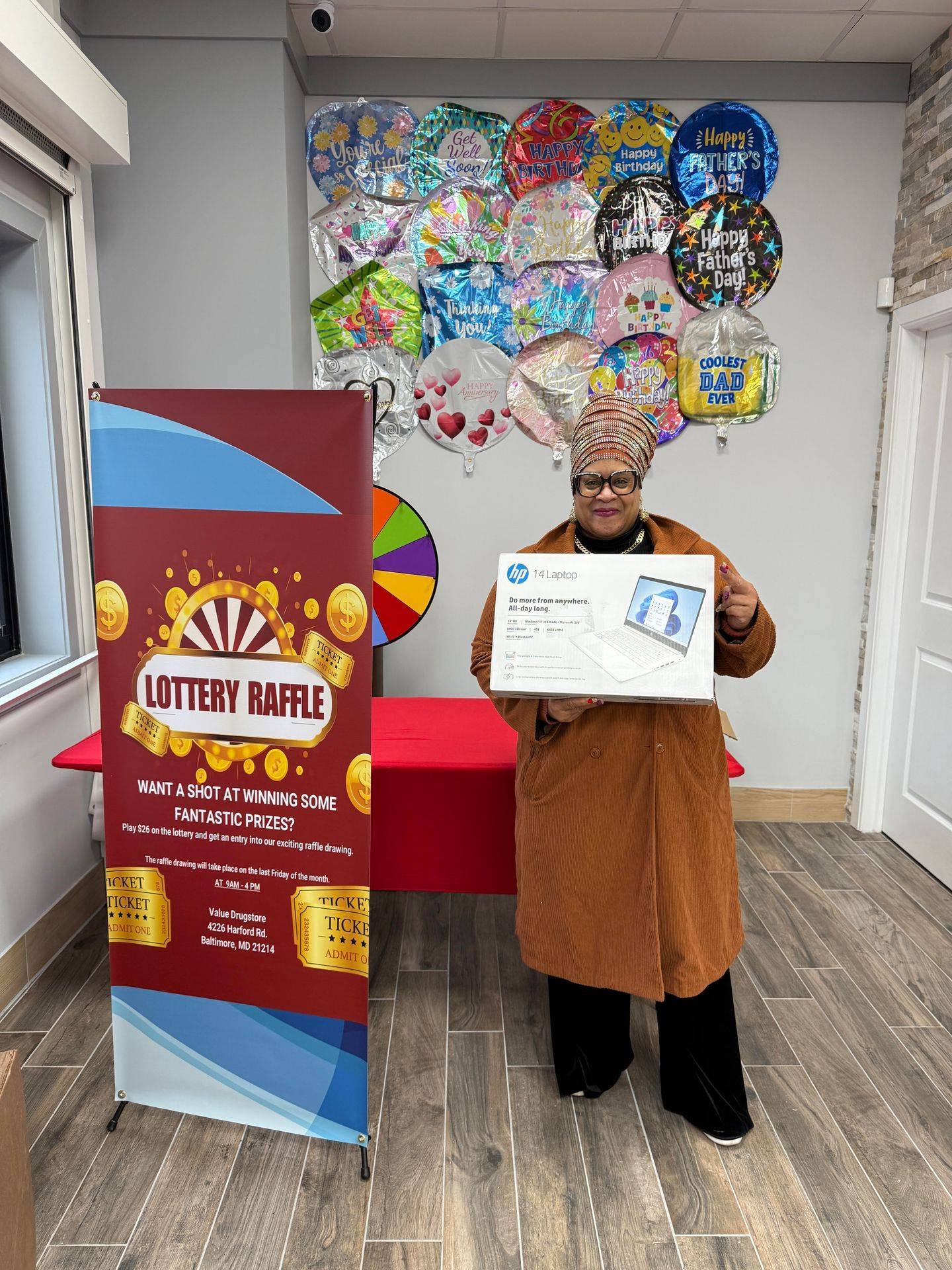 Woman holding an HP laptop in front of a lottery raffle banner at Value Drugstore, with colorful balloons in the background celebrating various occasions.